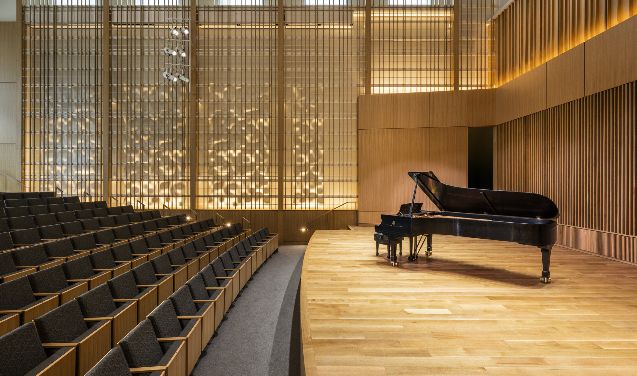 Angled view from side of stage looking toward wood design on wall illuminated from behind. Grey theater seats with light wood