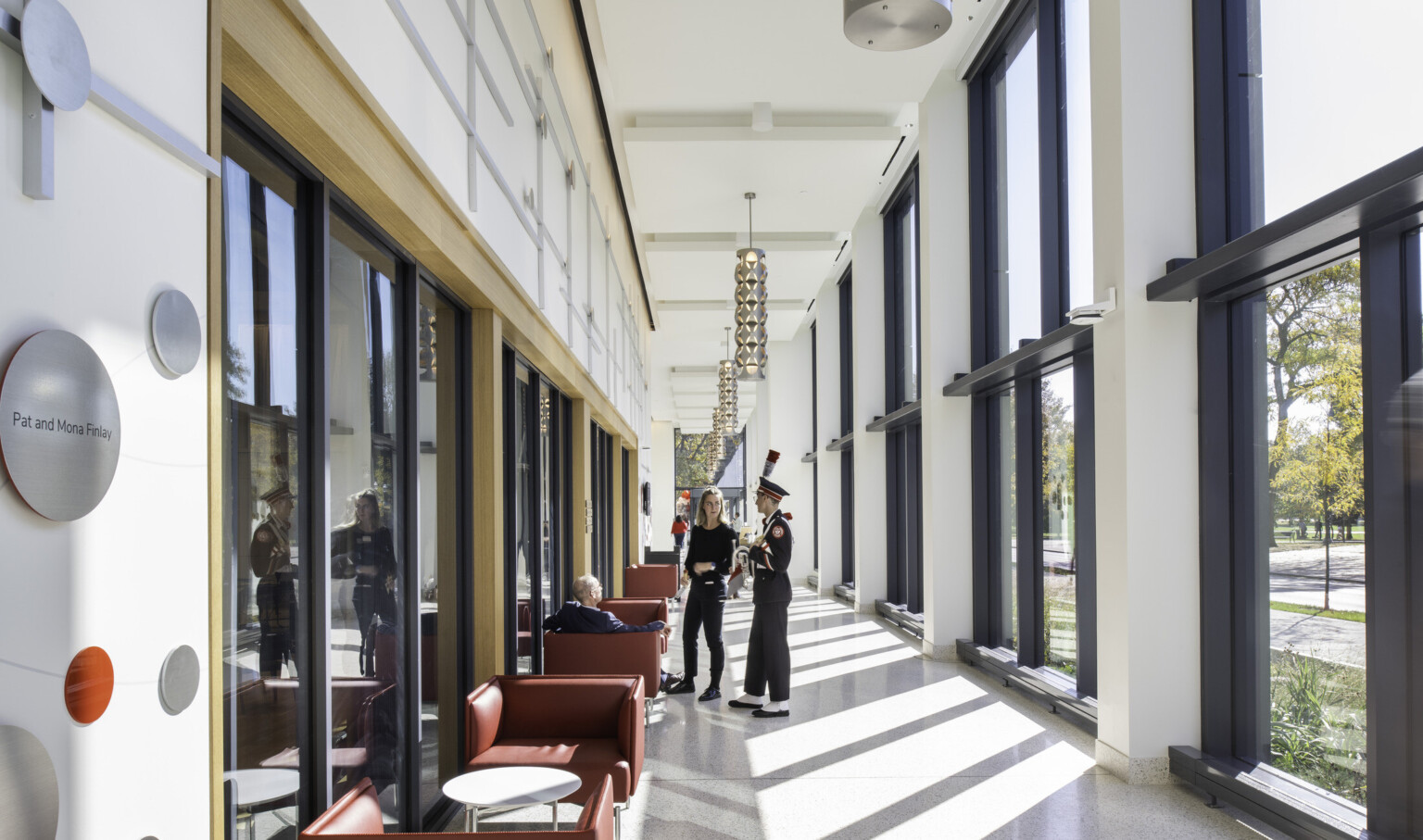 View of double height hallway with floor to ceiling windows to courtyard with red seating, metal pendant lights, and students conversing View of double height hallway with floor to ceiling windows to courtyard with red seating, metal pendant lights, and students conversing