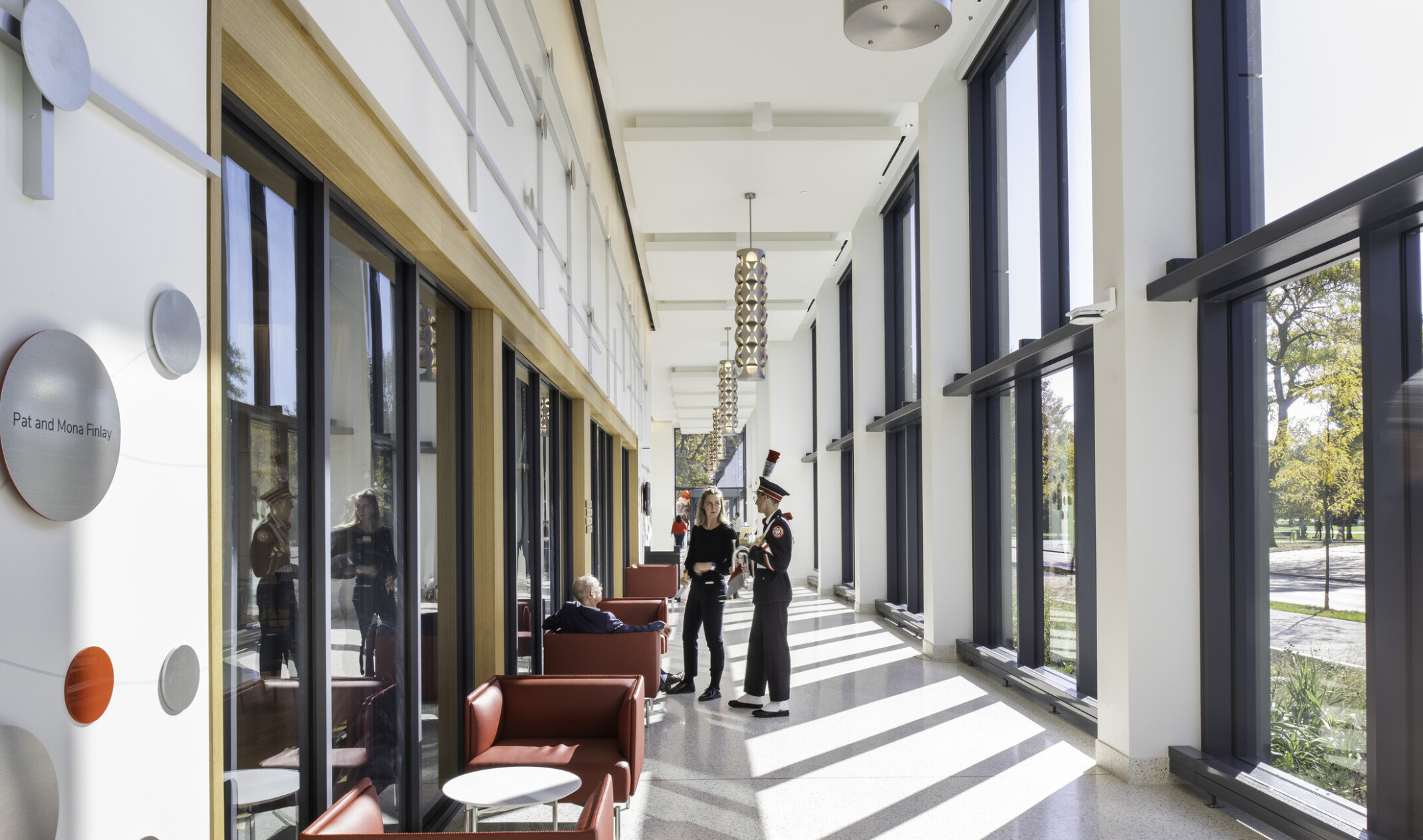 View of double height hallway with floor to ceiling windows to courtyard with red seating, metal pendant lights, and students conversing