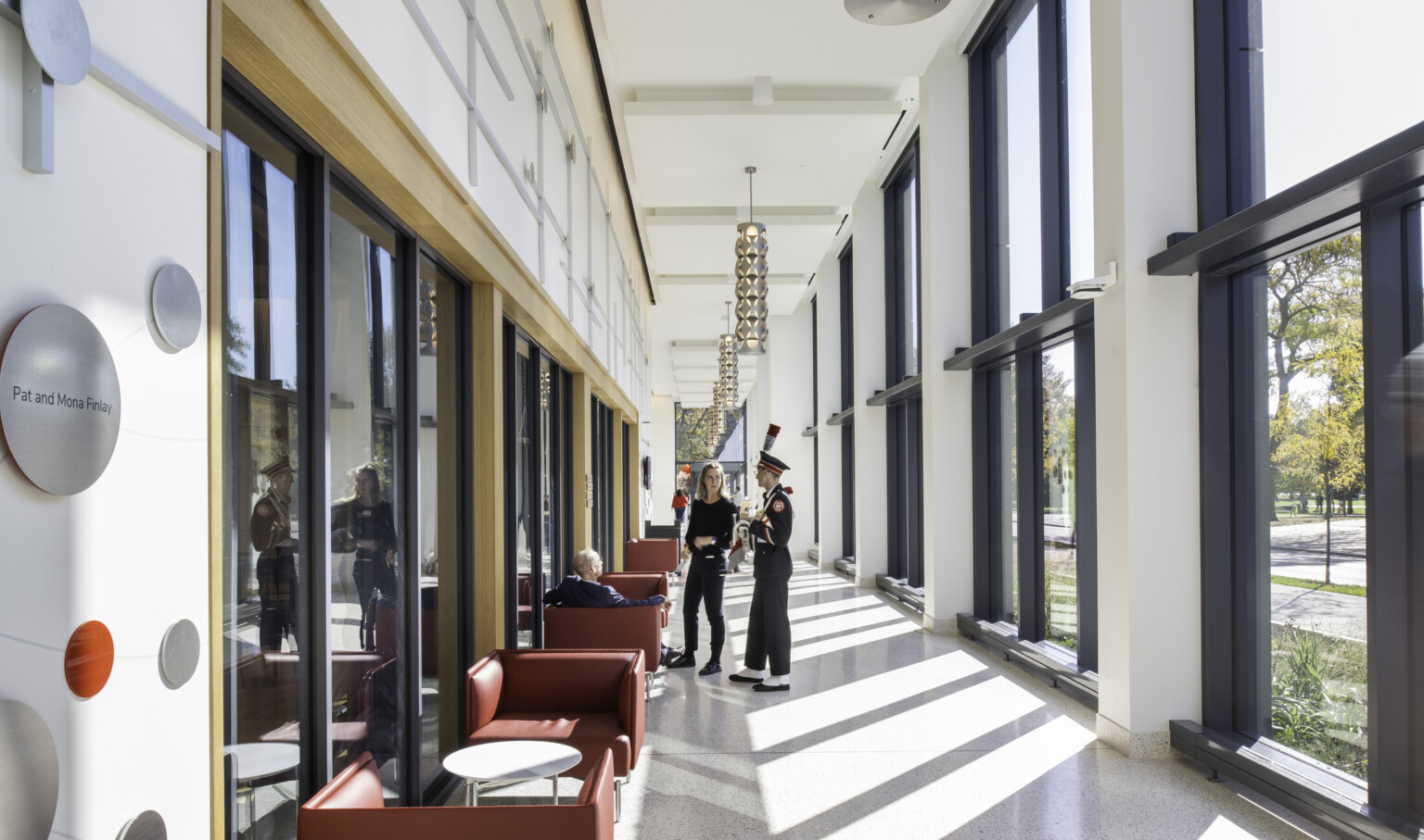View of double height hallway with floor to ceiling windows to courtyard with red seating, metal pendant lights, and students conversing