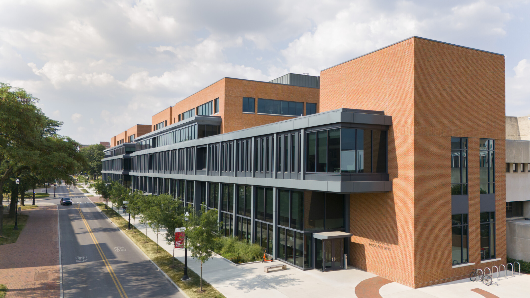 A modern academic building with brick and glass exterior, featuring large windows and a sidewalk lined with trees and lampposts.