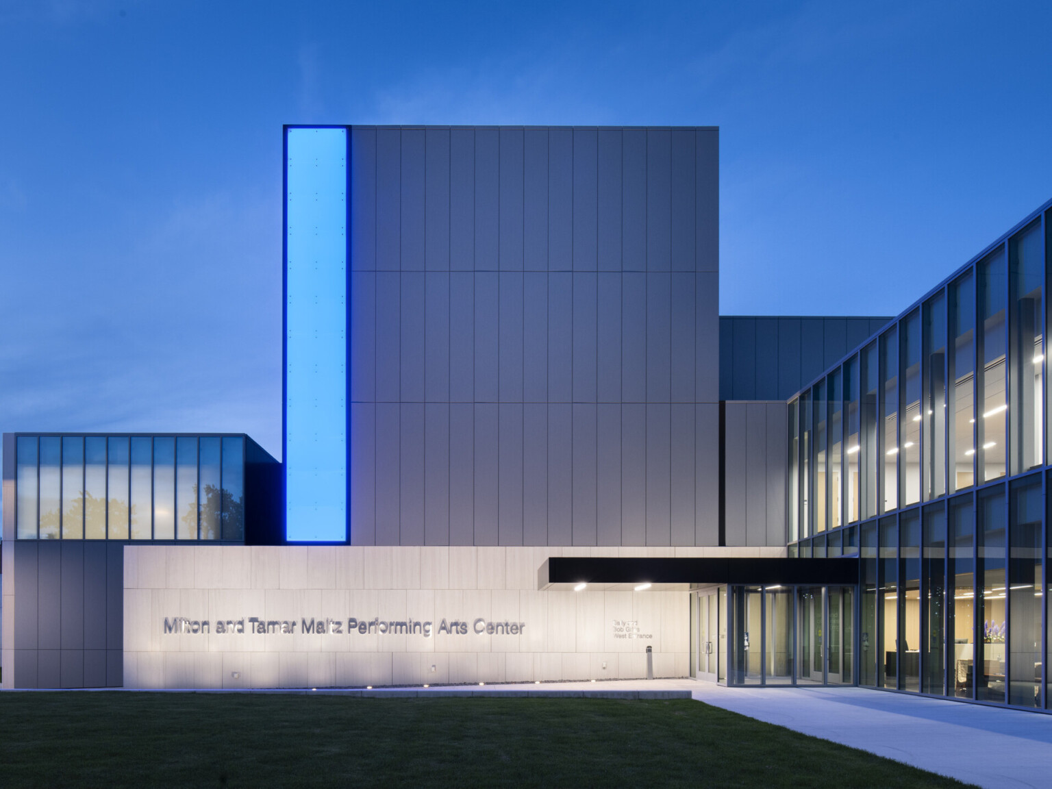 Side view of entry facing concrete wall and sign, grey building extends three stories above with a blue light panel on left Side view of entry facing concrete wall and sign, grey building extends three stories above with a blue light panel on left