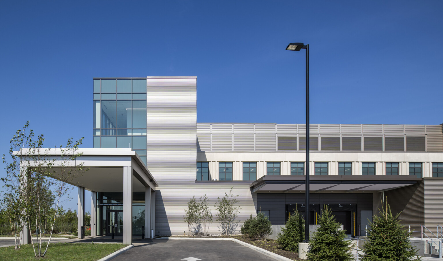 Exterior of outpatient care facility entrance, grey steel cladding facade, double-height and floor-to-ceiling windows brings sunlight indoors, parking lot and outdoor lighting, overhang at entrance protects folks from inclement weather