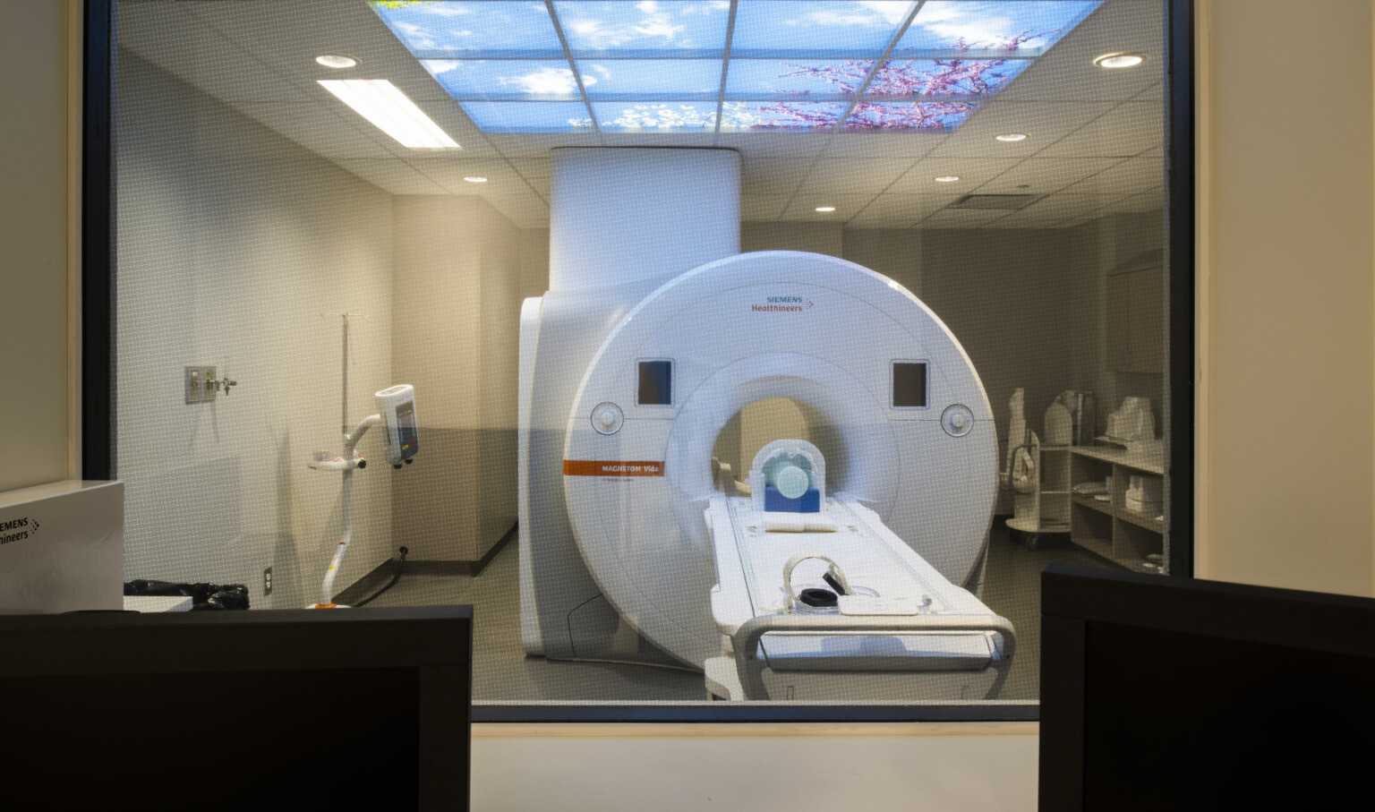 View through interior window to CAT Scan machine in white room with fake skylight showing blue sky with white fluffy clouds
