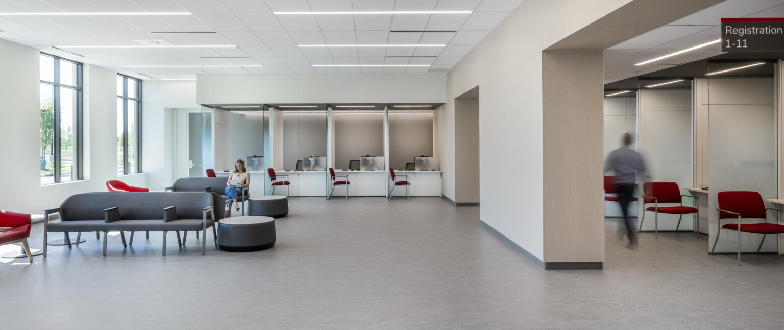 White waiting room with wall separating seating area with grey chairs from registration area with red chairs and glass partitions