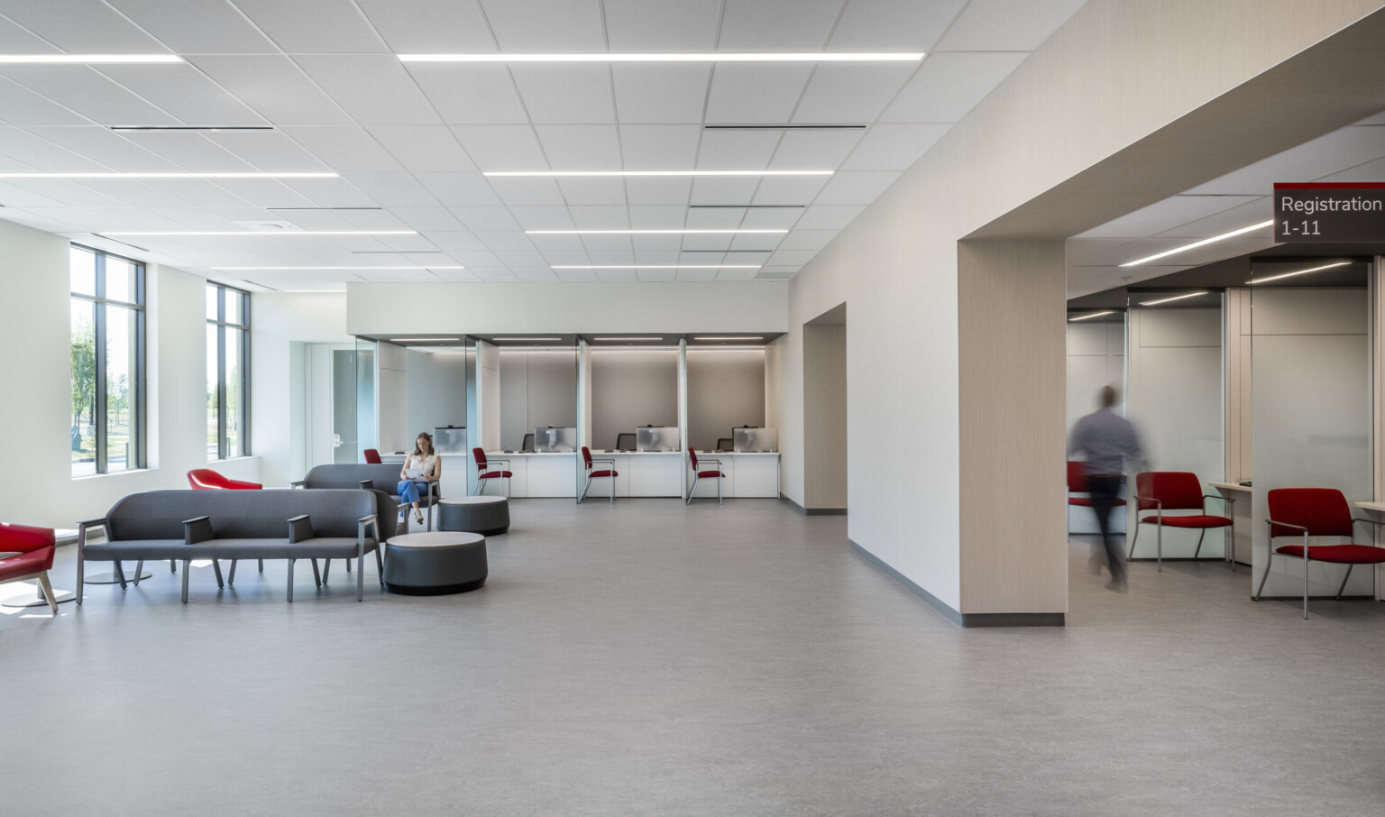 White waiting room with wall separating seating area with grey chairs from registration area with red chairs and glass partitions