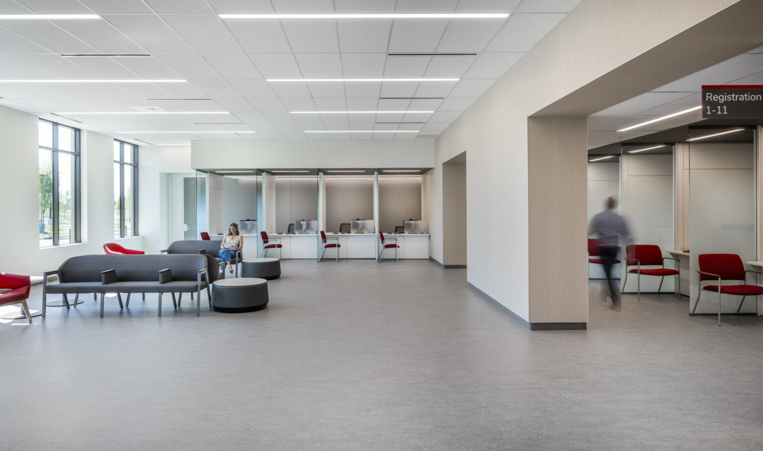 White waiting room with wall separating seating area with grey chairs from registration area with red chairs and glass partitions