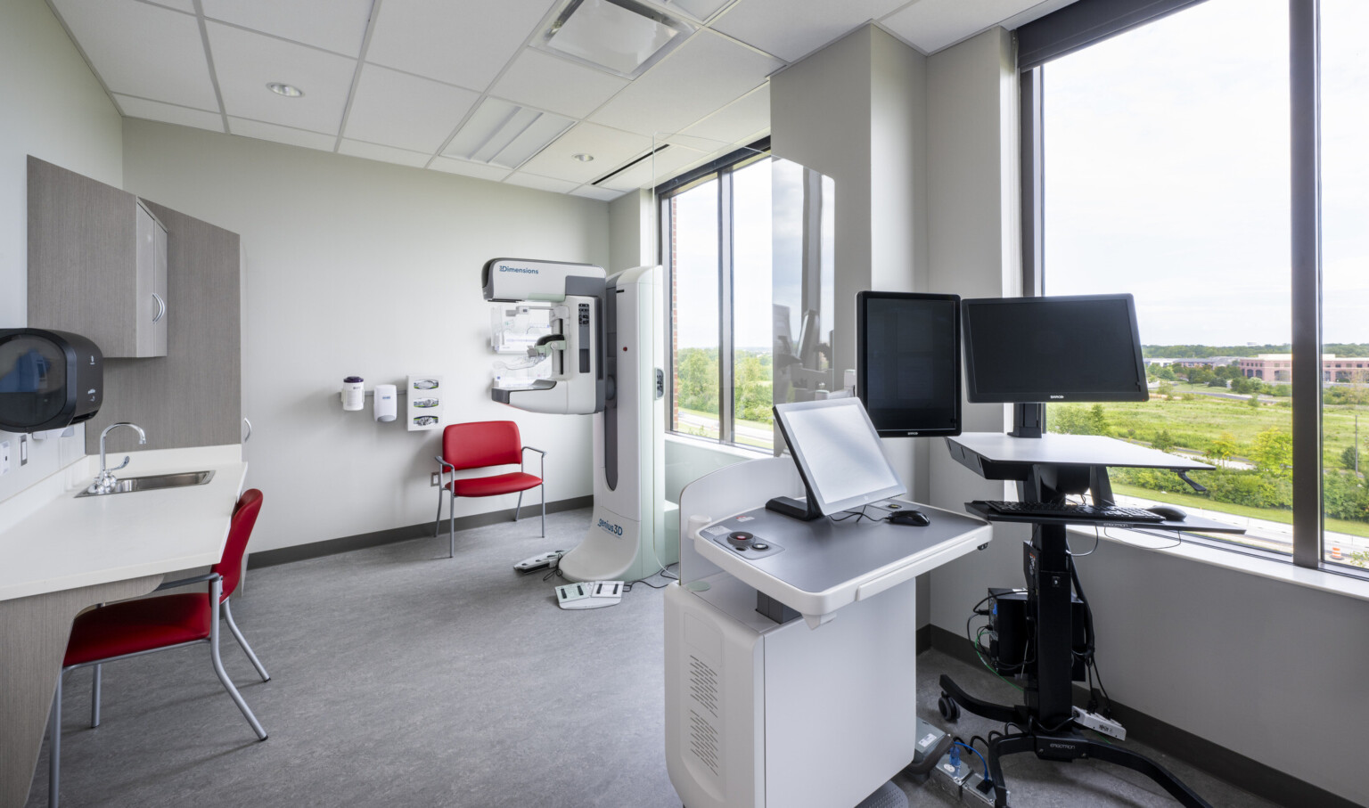 Patient care space with sink, portable standing desk with computer monitors, healthcare equipment. Large windows on right wall
