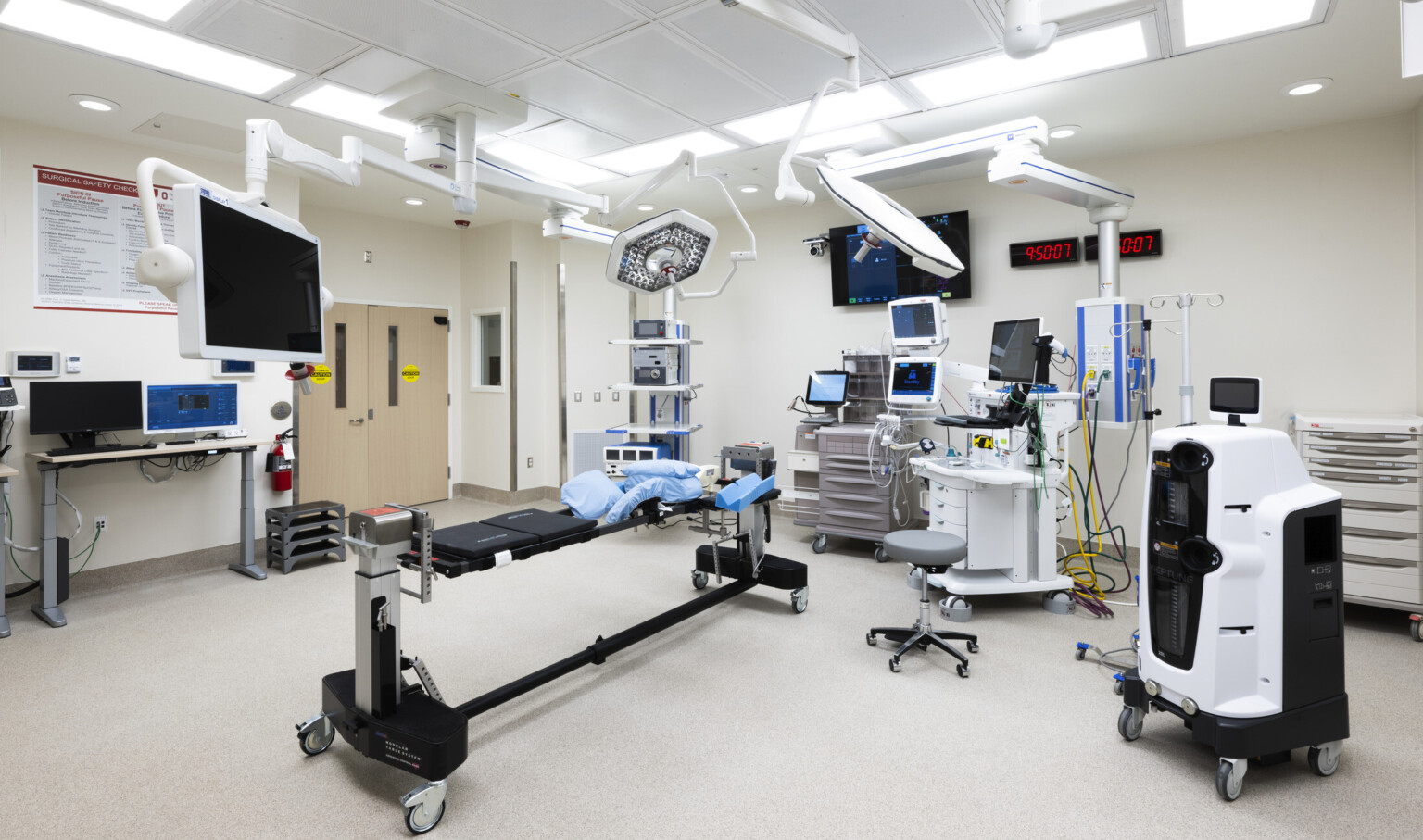Operating room with medical equipment along outside edge of white room with two digital wall clocks. Equpment hangs above
