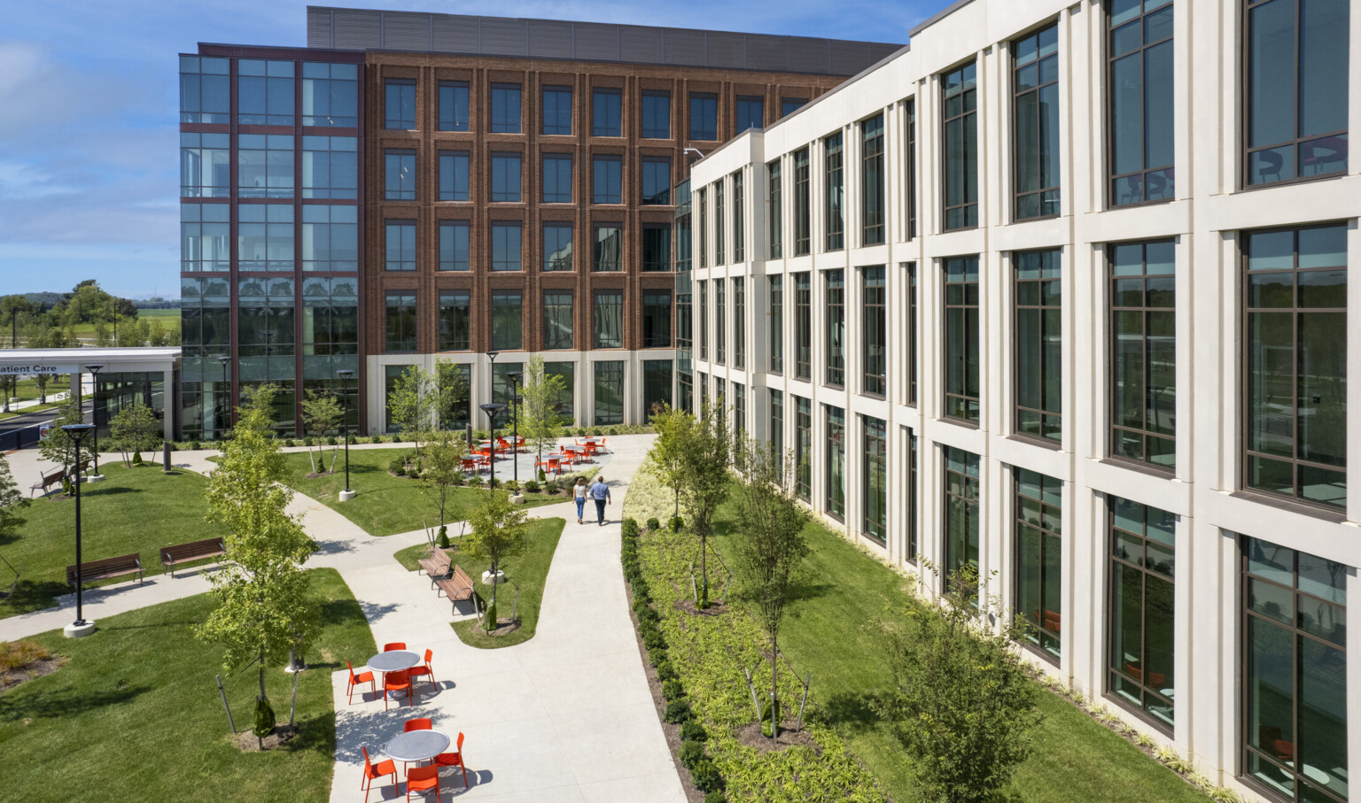 Aerial view of a concrete pathway along a white building surrounded by lush landscaping. Seating areas with grey table and orange chairs along with path