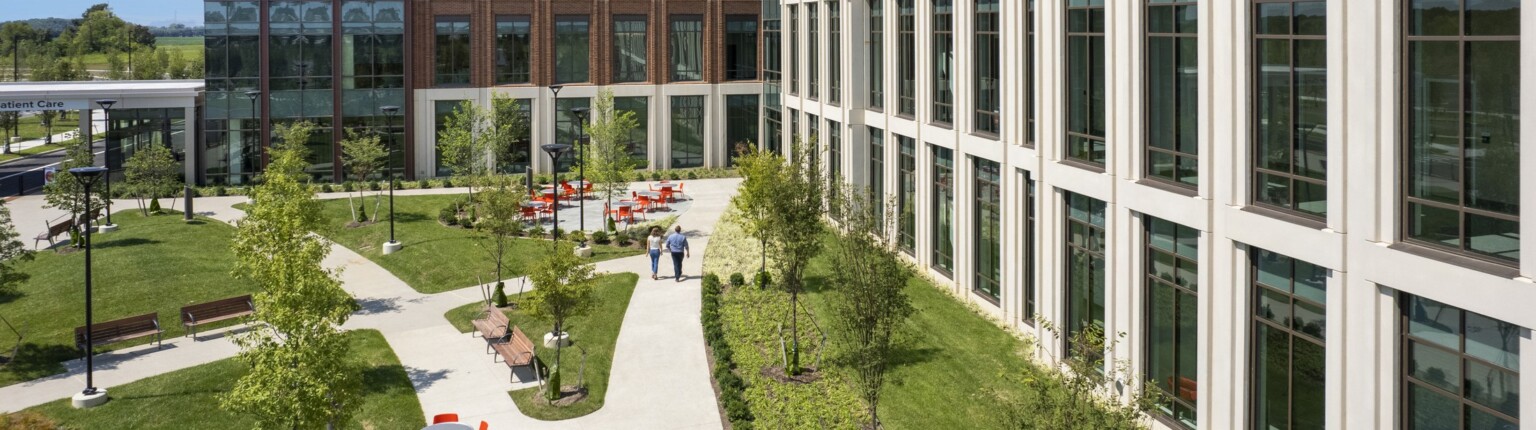 Aerial view of a concrete pathway along a white building surrounded by lush landscaping. Seating areas with grey table and orange chairs along with path