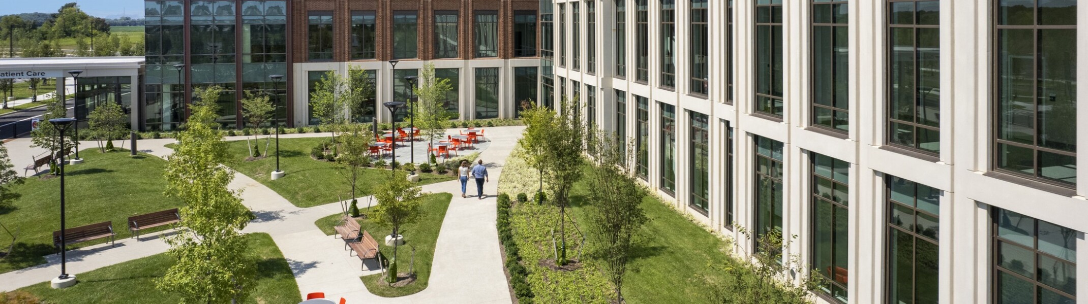 Aerial view of a concrete pathway along a white building surrounded by lush landscaping. Seating areas with grey table and orange chairs along with path
