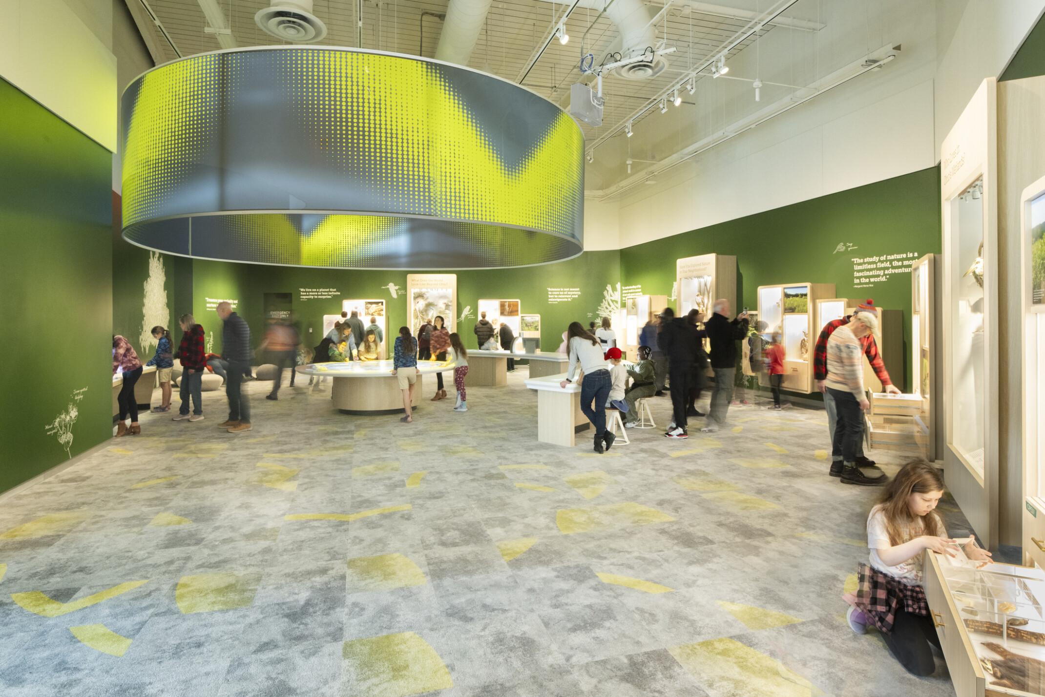 Visitors interacting with exhibits in a brightly lit indoor exhibition space with green walls and a large circular ceiling fixture