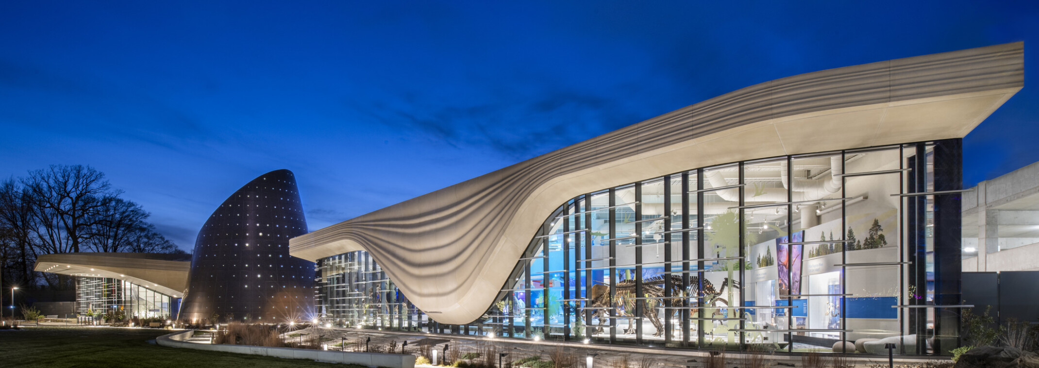 Nighttime image of a building with a white striped overhang that bows in center of right building section, an angled tube metal structure is at the building center