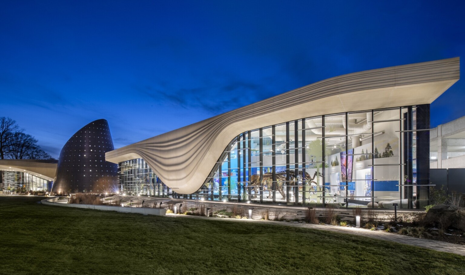 Nighttime image of a building with a white striped overhang that bows in center of right building section, an angled tube metal structure is at the building center