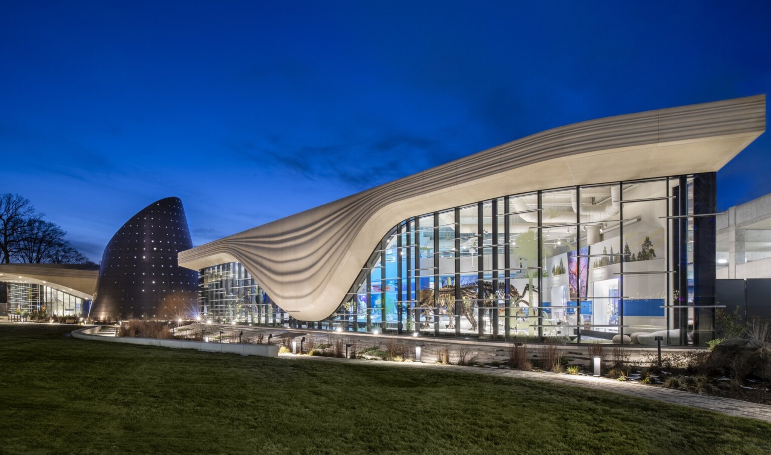 Nighttime image of a building with a white striped overhang that bows in center of right building section, an angled tube metal structure is at the building center