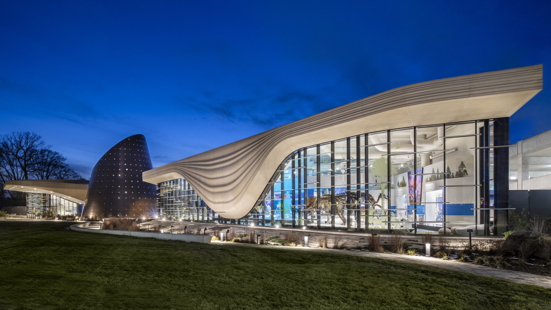 Nighttime image of a building with a white striped overhang that bows in center of right building section, an angled tube metal structure is at the building center