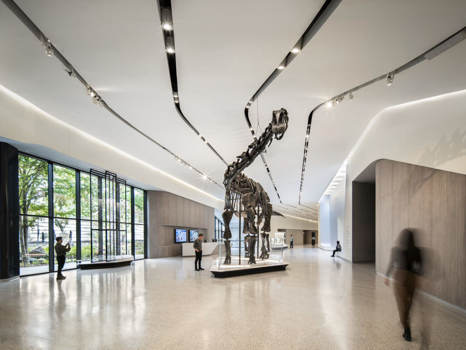 A dinosaur skeleton in an exhibition hall with people walking and viewing the exhibit in the large space with white walls, light tile flooring at the Cleveland Museum of Natural History