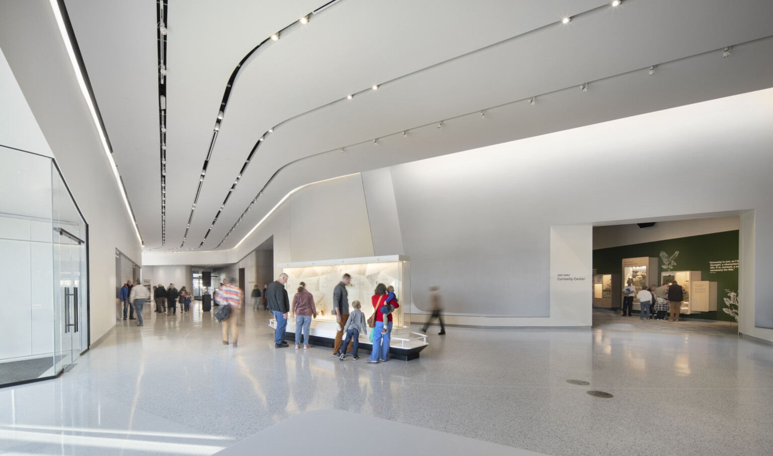 A spacious, modern museum lobby with a sleek, minimalist design featuring curved white walls and a polished terrazzo floor. Several people are walking or standing near a central display case, while others move toward different exhibit entrances. Recessed ceiling lights create a bright, evenly lit environment. On the right, an open doorway leads to a green-walled gallery space.