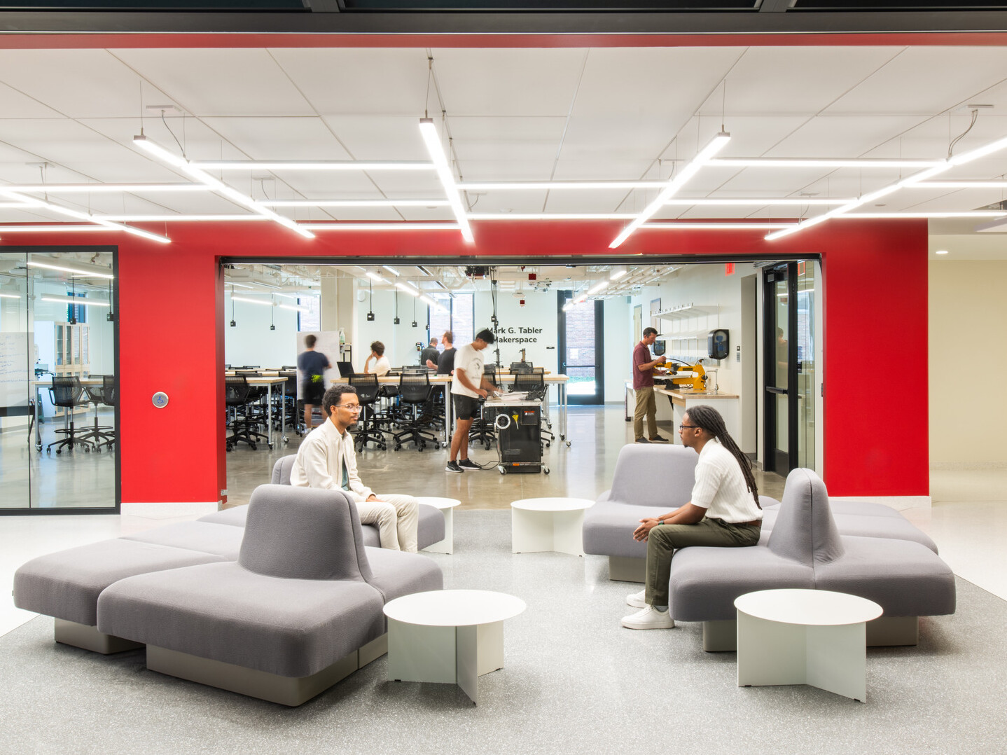 modern community space in a science building; comfortable modular, sitting space looks into an open concept lab behind floor to ceiling glass