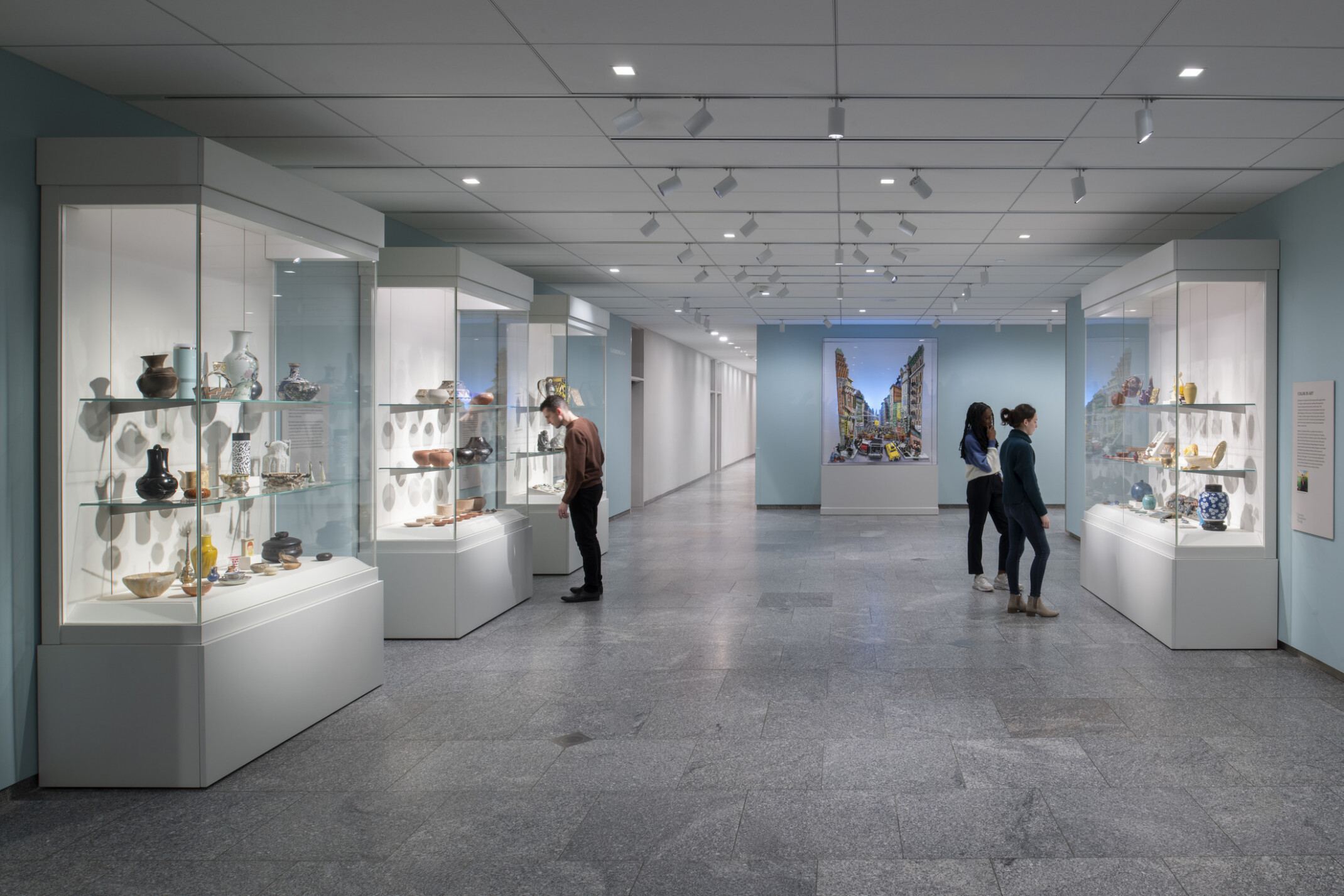 Museum hallway with glass cases of ceramic art and three visitors, ending with a colorful butterfly artwork.
