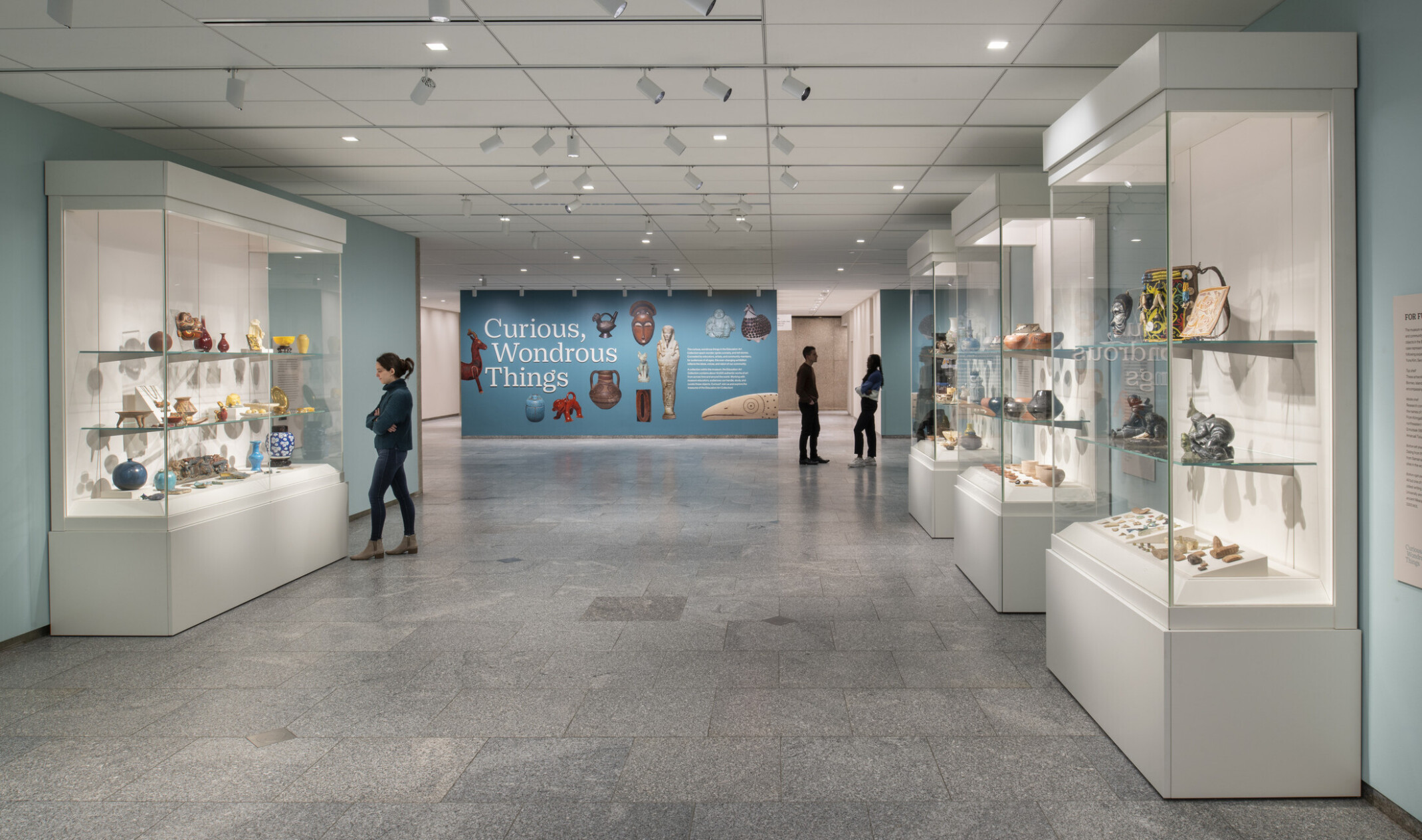 Museum hallway with glass cases of ceramic art and three visitors, ending with a colorful butterfly artwork.