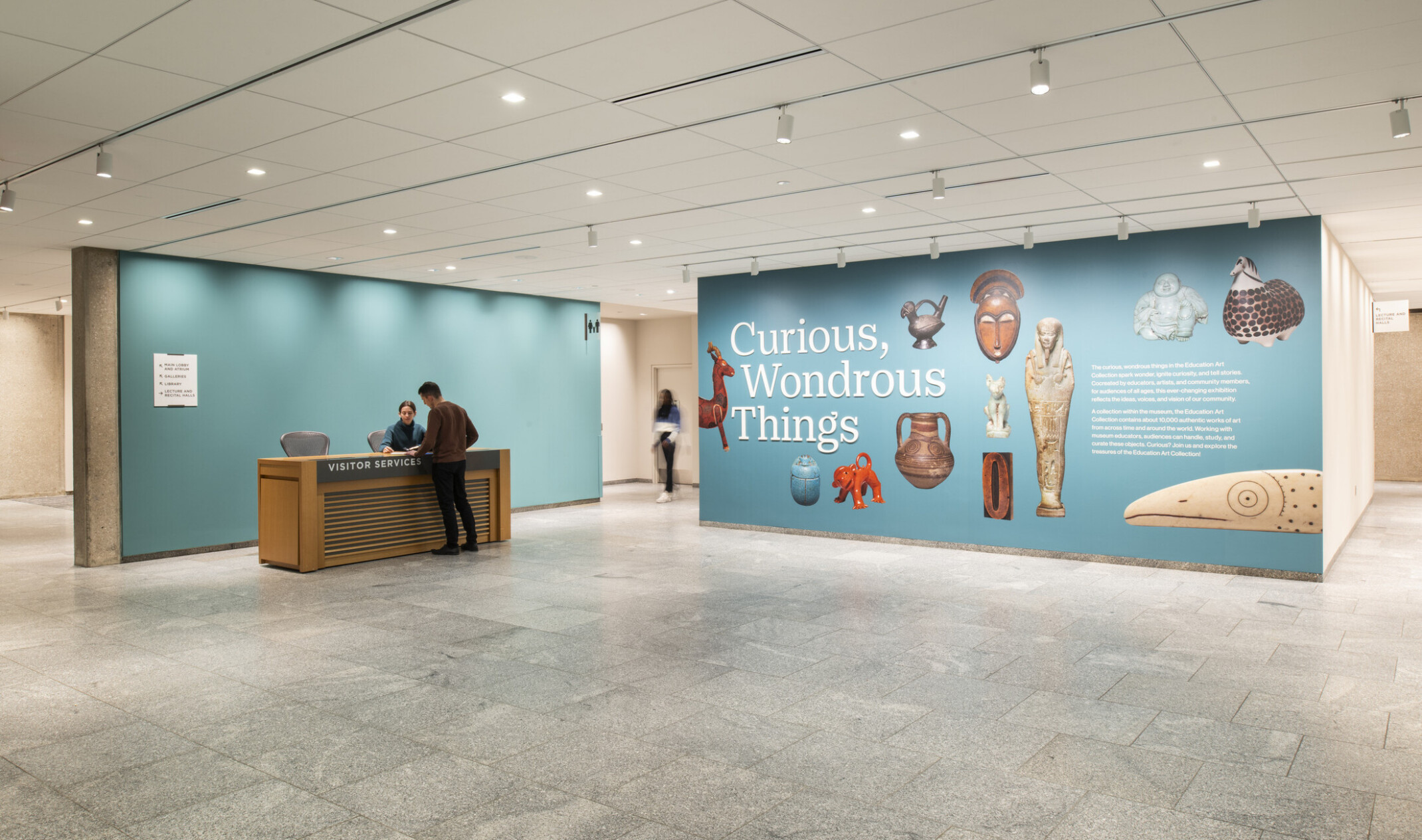 Gallery entrance with reception desk and a teal wall displaying ‘Curious, Wondrous Things’ surrounded by artifact images.