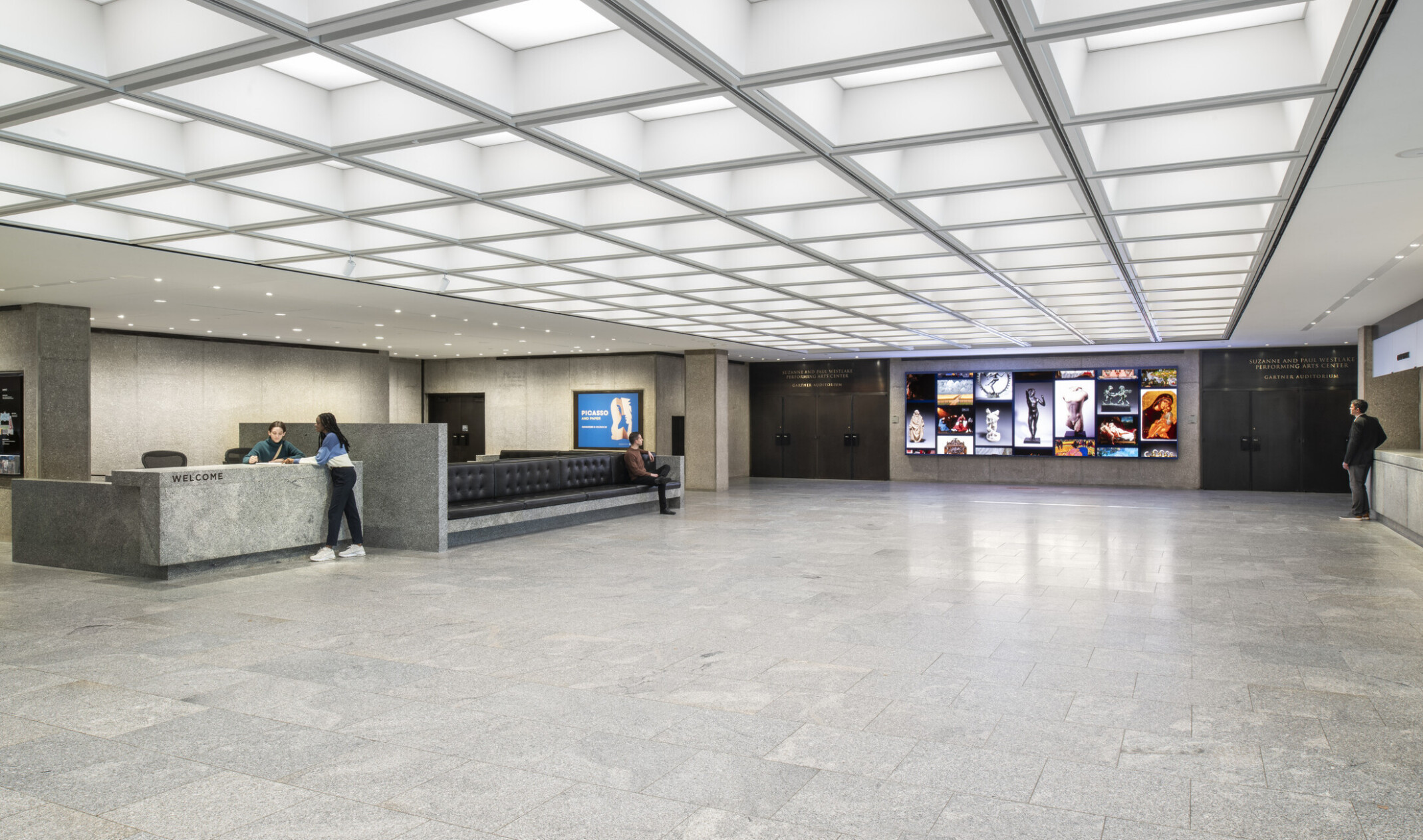 Modern lobby with gray stone floors, reception desks on both sides, central seating, and large digital screens on the back wall.