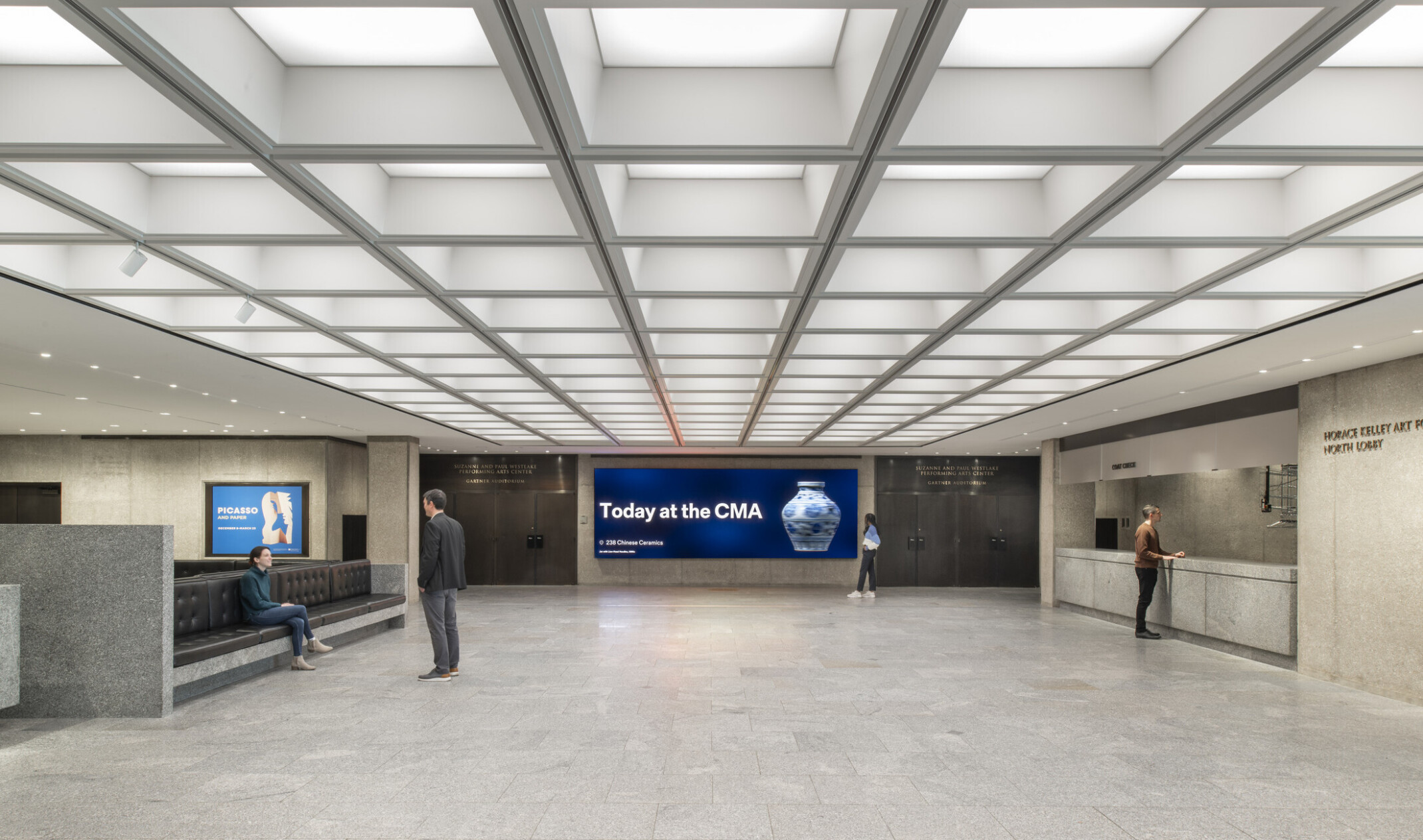 Modern lobby with gray stone floors, reception desks on both sides, central seating, and large digital screens on the back wall.