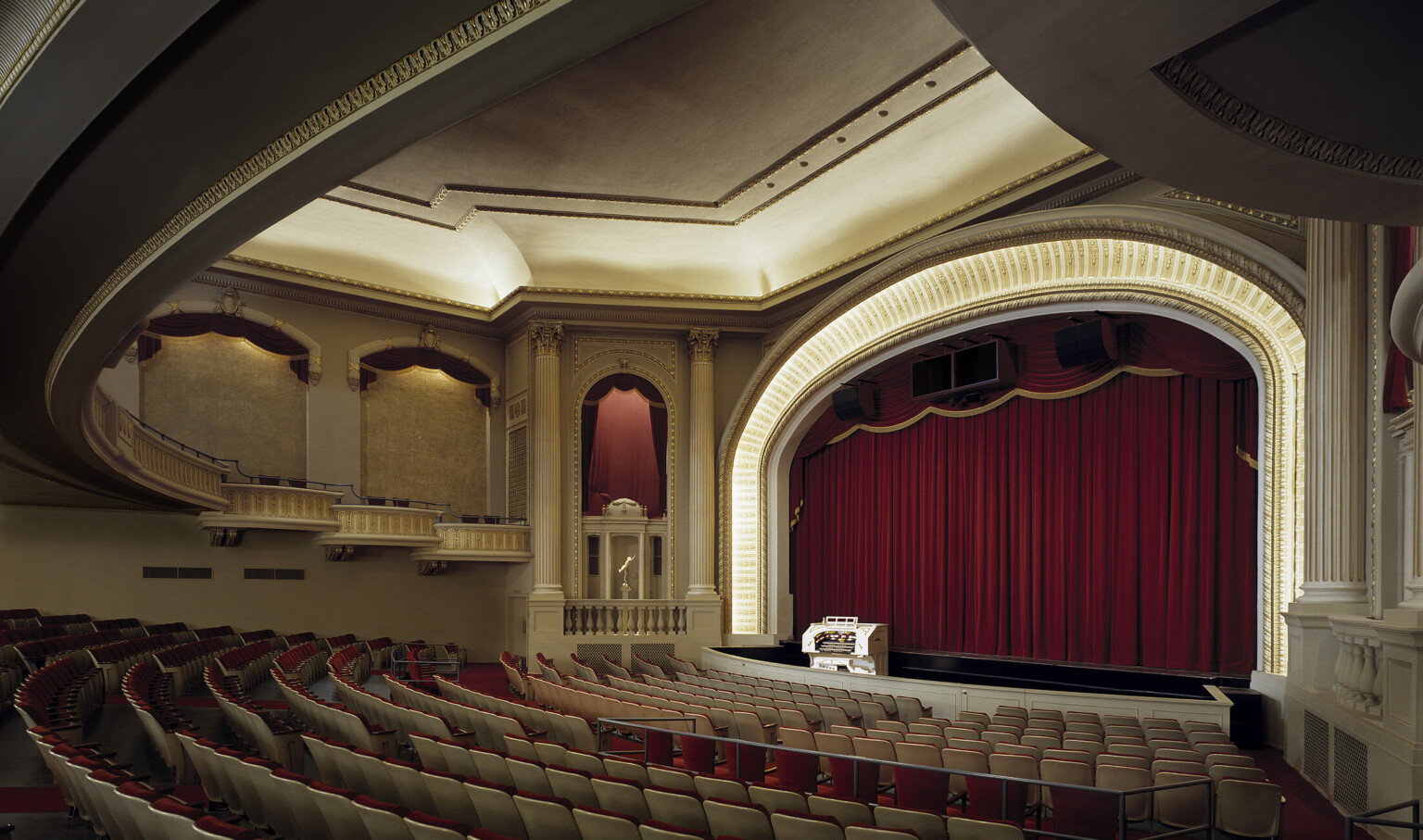 historic 1927 vintage grand theater with curved lowered ceiling design, dramatic lighting, red curtain behind stage, orchestra pit, traditional red theater seating