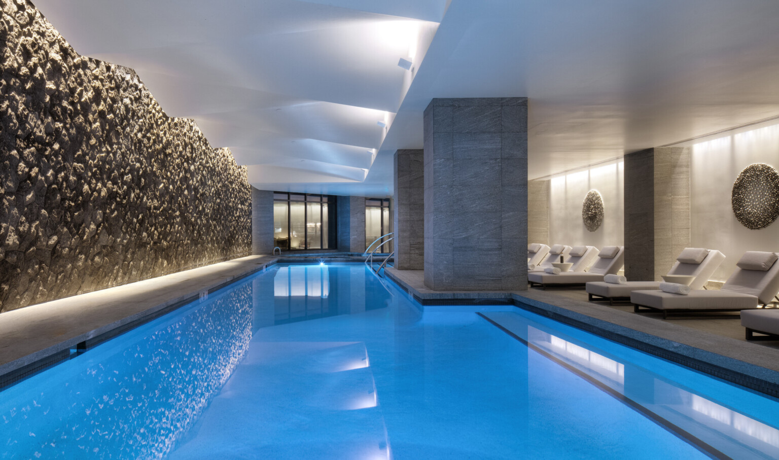 A hotel indoor pool lined with cushioned chaise loungers along a swimming pool lit up blue