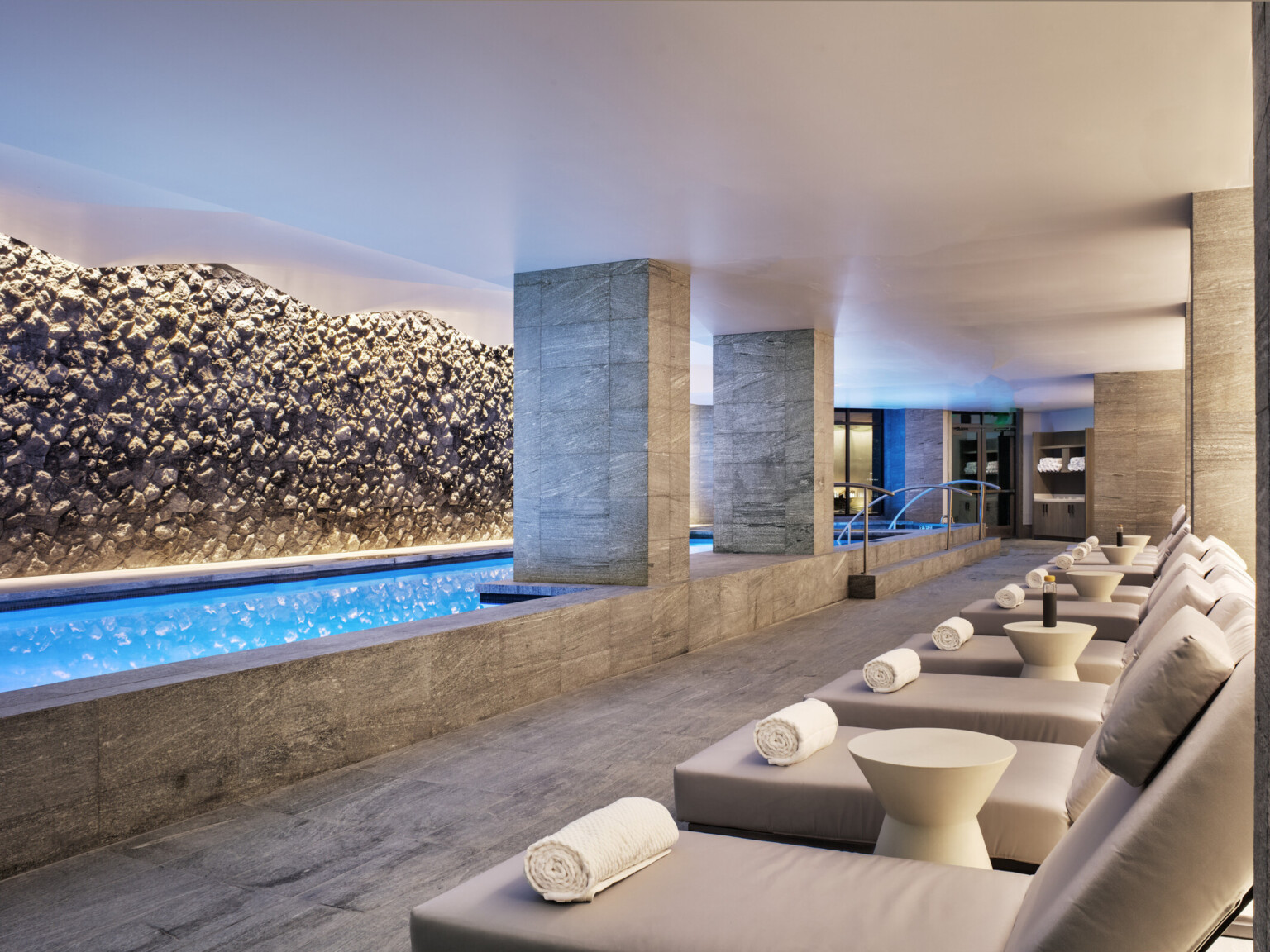 An upscale indoor pool lined with cushioned chaise loungers looking at a swimming pool lit up blue