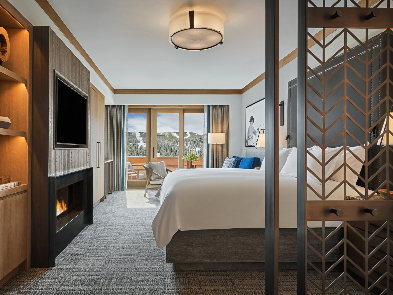 Modern hotel room with a large bed, balcony view of snowy mountains, and a fireplace under a mounted TV.