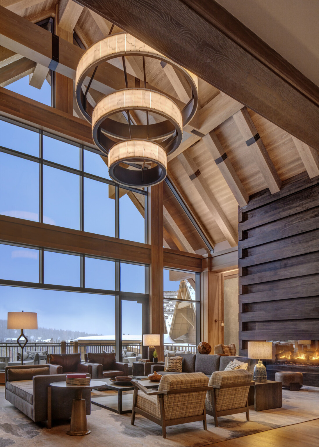 Spacious hotel seating area with vaulted wooden ceiling, large windows, and a chandelier hanging in the middle of the room with a seating area in from of a fireplace below