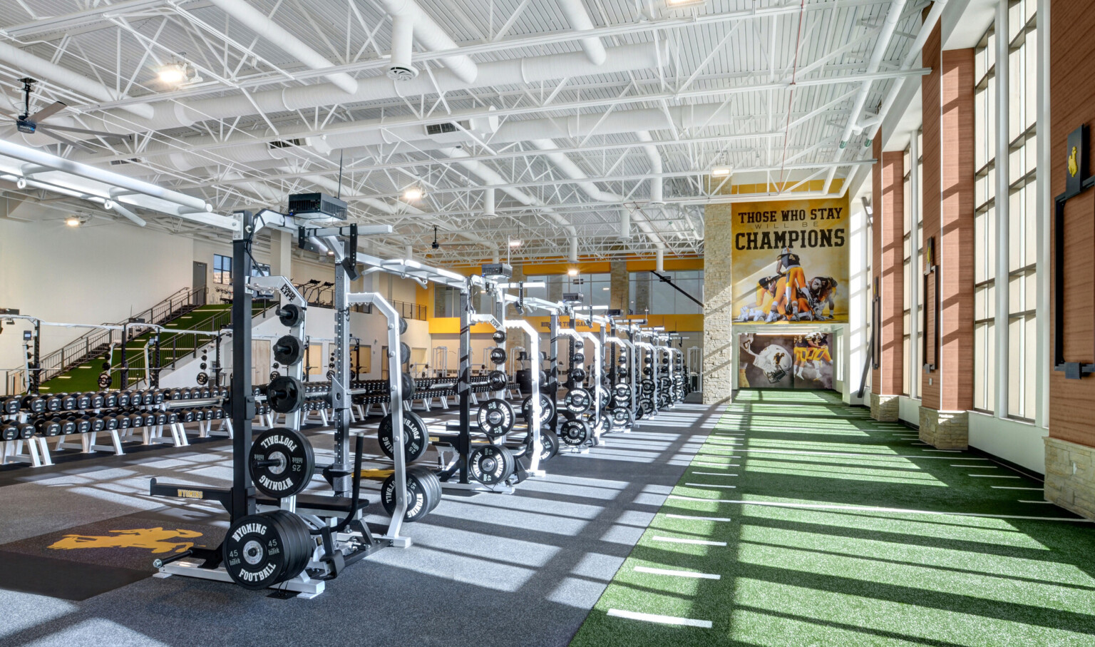 Turf floor with yard marks along double height windows. Ahead, a mural reading Those Who Stay Will Be Champions. Weights left