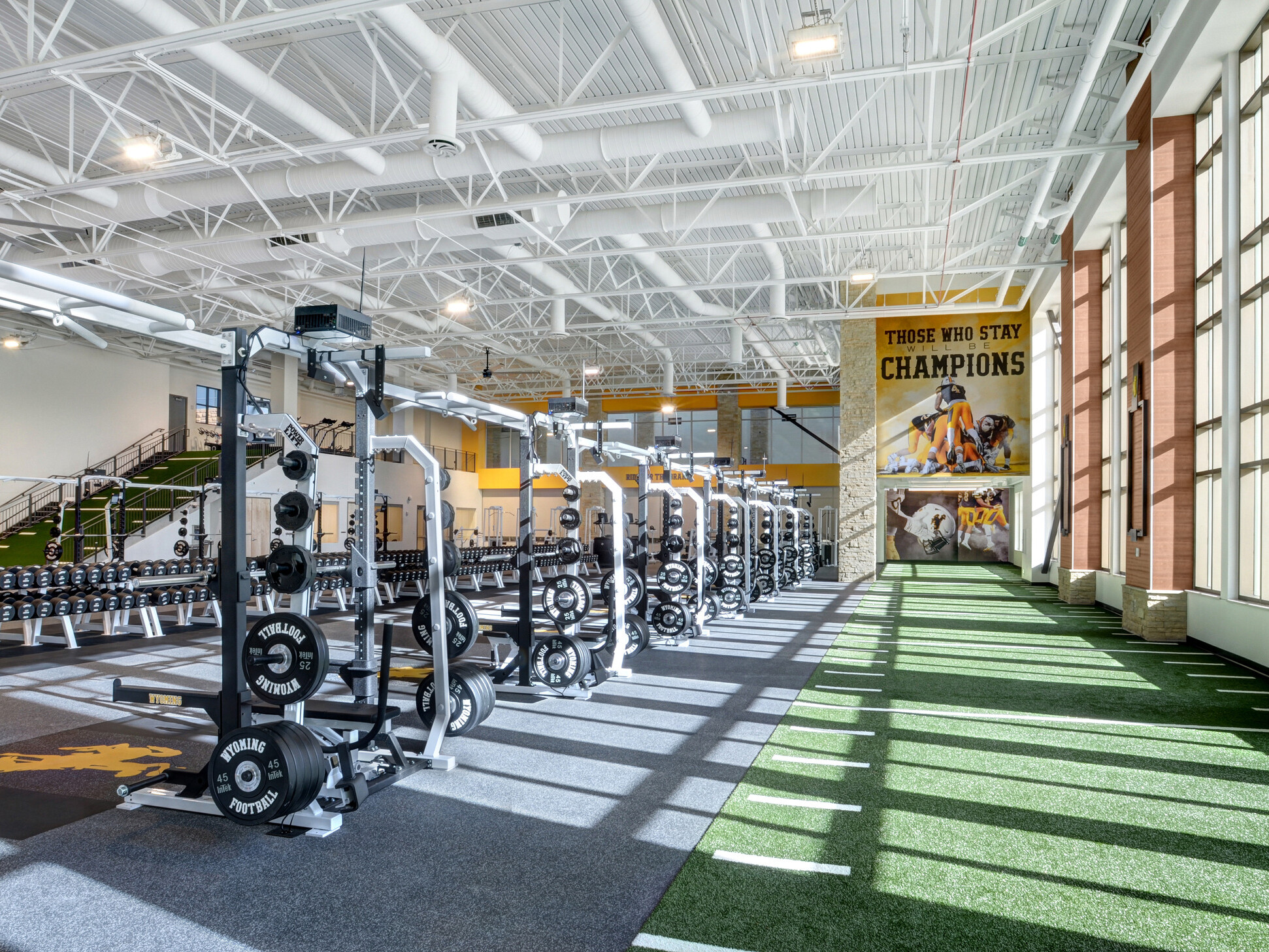 Turf floor with yard marks along double height windows. Ahead, a mural reading Those Who Stay Will Be Champions. Weights left