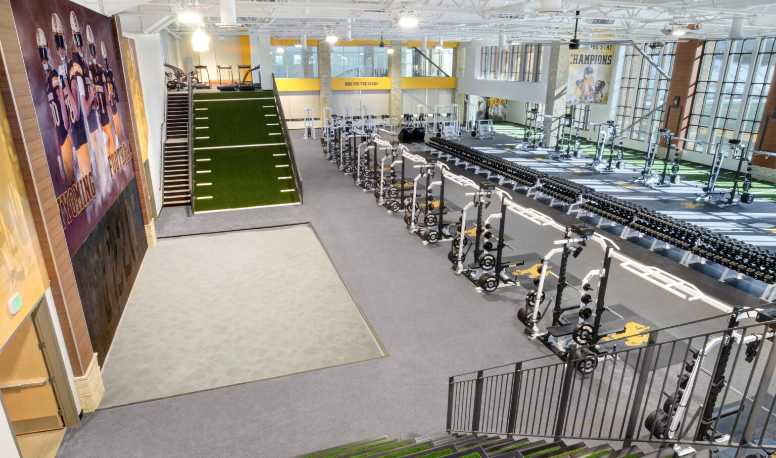 Double height training facility with rows of equipment. Left, a Wyoming Football mural on wall beside stairs and turf slope