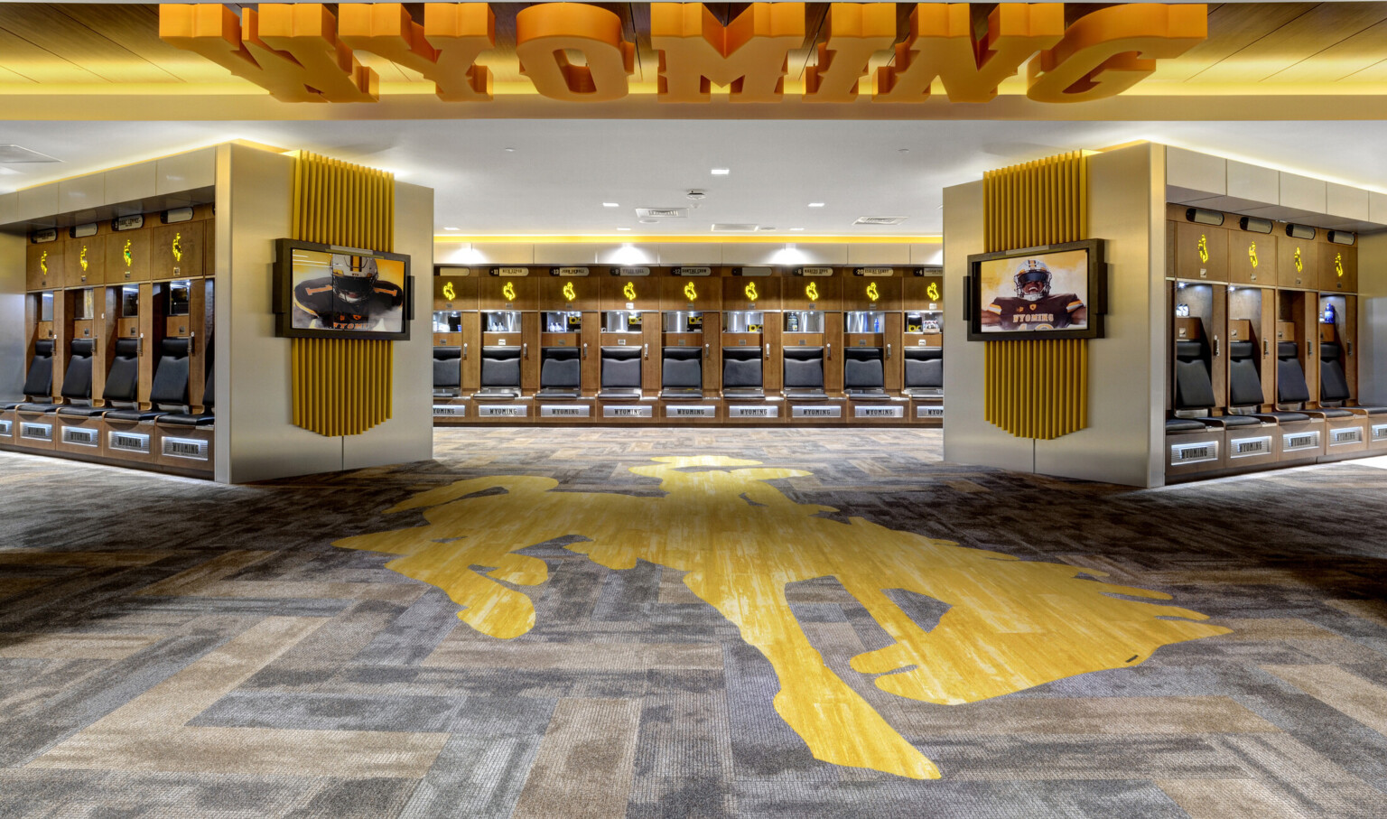 Locker room with yellow accents. Mascot illuminated above wood lockers. Yellow Wyoming sign on ceiling and mascot on carpet