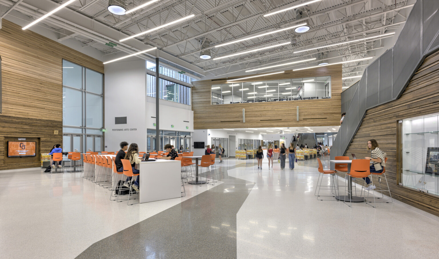 Large open common area; light gray floors and paneled wood accent walls offset the bright orange chairs for school children to work on laptops; staircase on the right leads down to the main space; Background upper level windows showcase another classroom