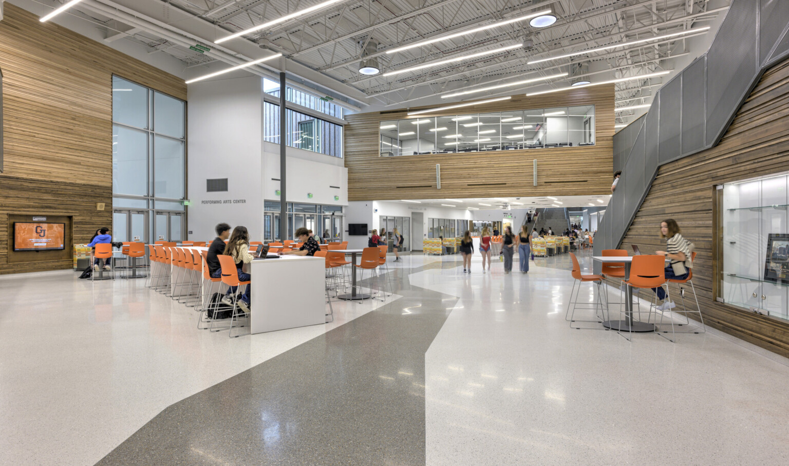Large open common area; light gray floors and paneled wood accent walls offset the bright orange chairs for school children to work on laptops; staircase on the right leads down to the main space; Background upper level windows showcase another classroom