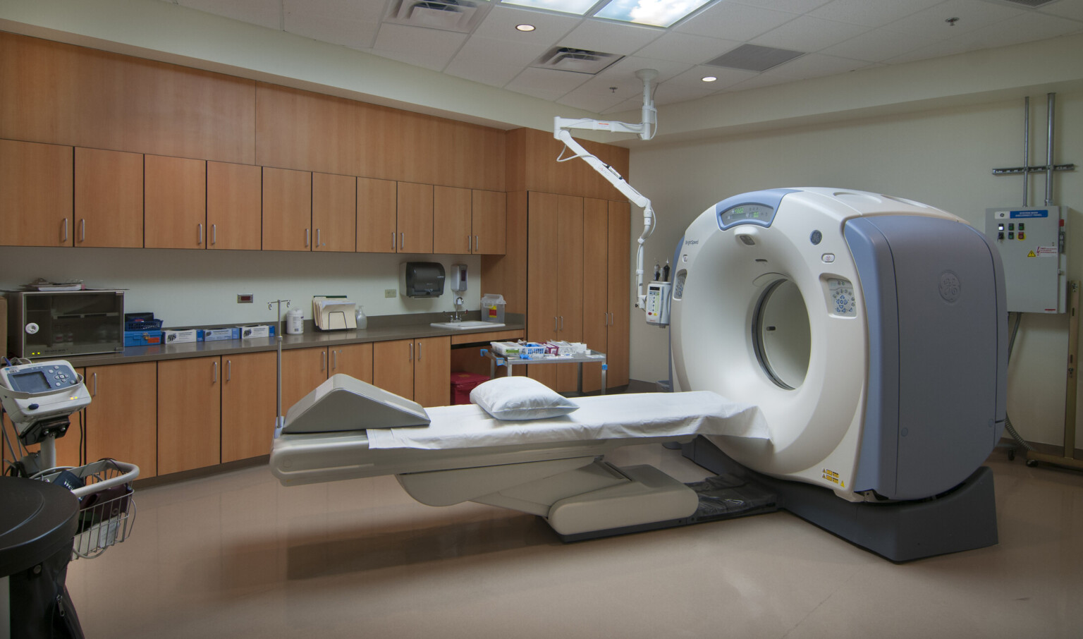 MRI machine in center of white room with wood cabinets right of shelves above and below counter space with sink