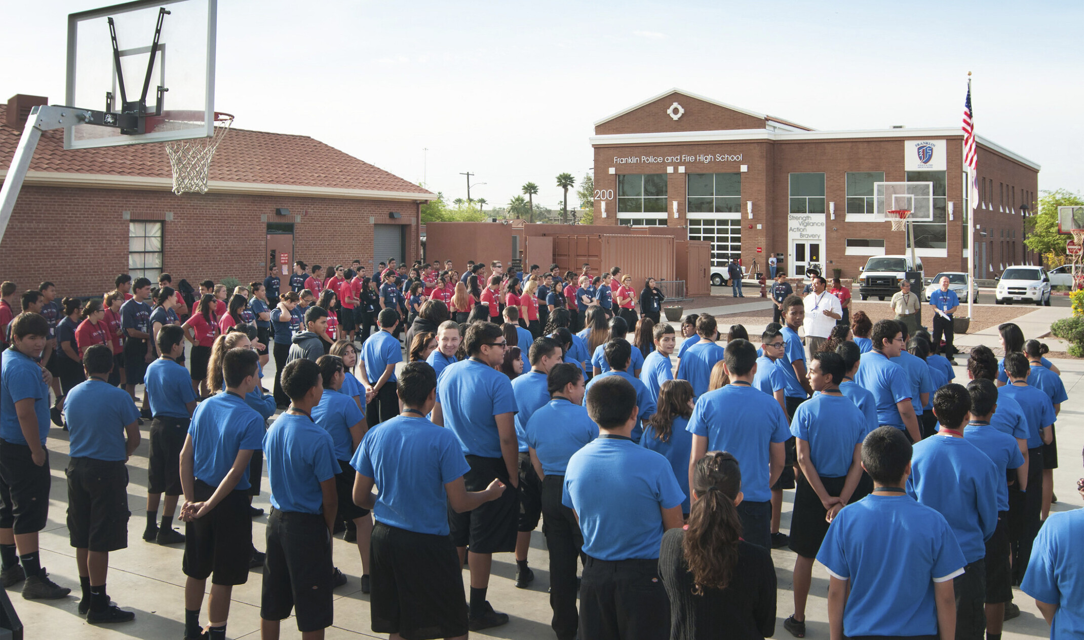 students in an assembly outside on the basketball courts in front of Franklin Police and Fire High School, 2 brick buildings