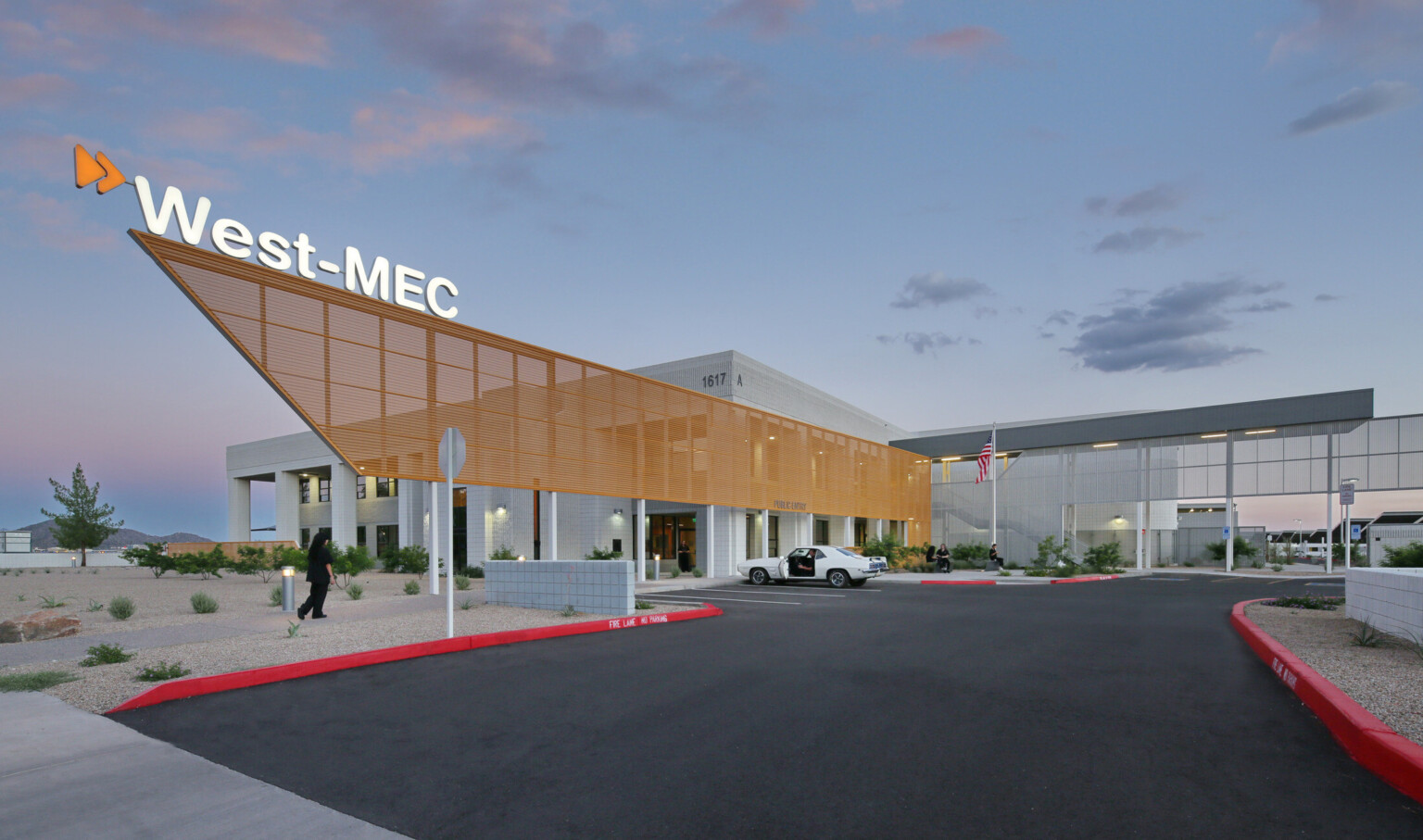 West-MEC Northeast Campus entrance with illuminated sign on top of orange perforated sunscreen extending past entrance West-MEC Northeast Campus entrance with illuminated sign on top of orange perforated sunscreen extending past entrance