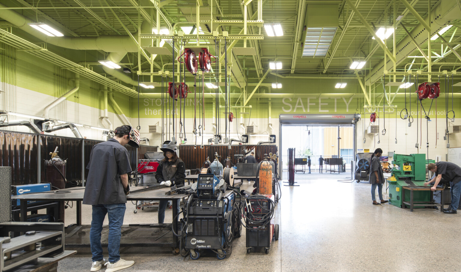 Students in a metal shop class with welding machines and cables hanging from the ceiling