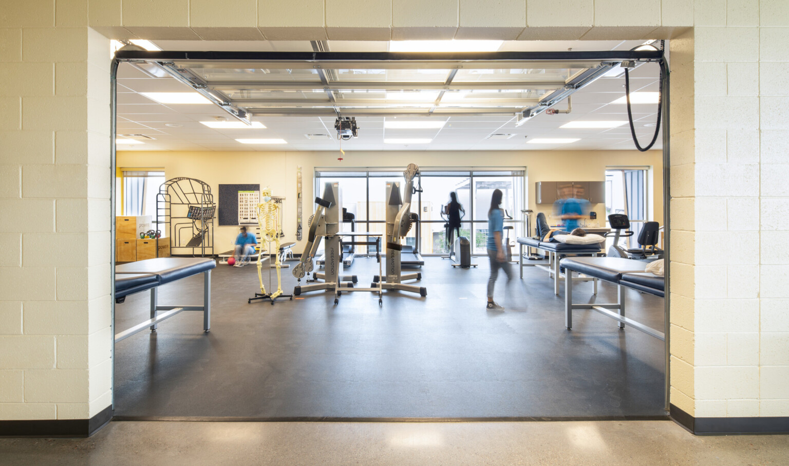 Inside a physical therapy room, a door opens to reveal a gym bustling with activity and exam tables
