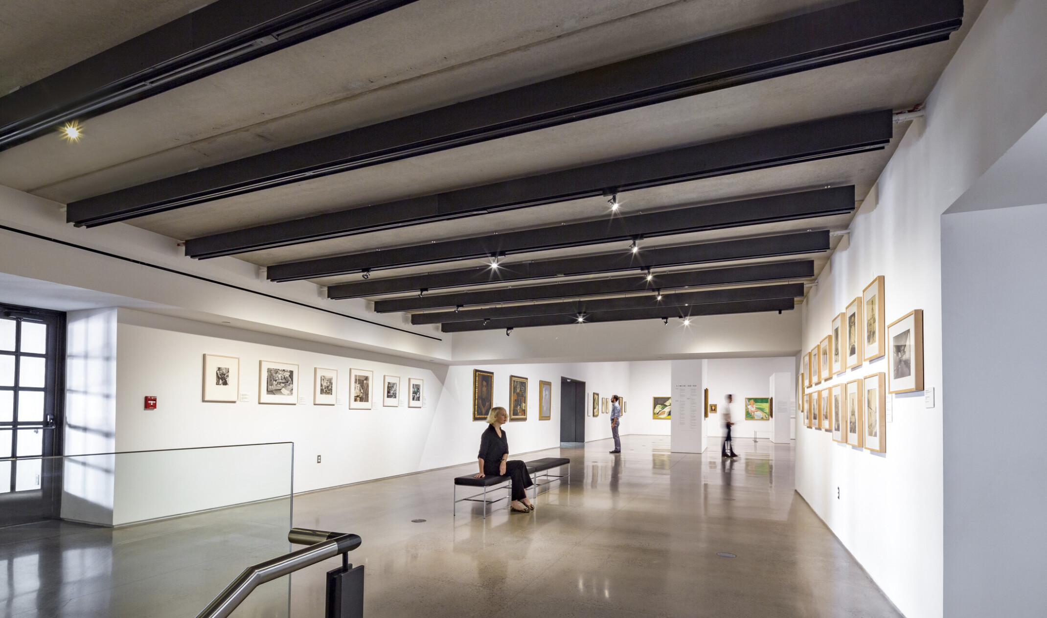 White gallery hall with paintings on the walls and benches along the center. Black beams cross the room's width with lighting