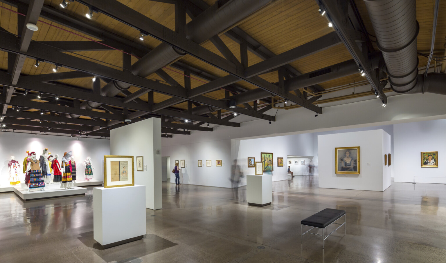 A gallery with paintings and a display of dressed mannequins, under a wood ceiling with exposed black support beams and pipes A gallery with paintings and a display of dressed mannequins, under a wood ceiling with exposed black support beams and pipes