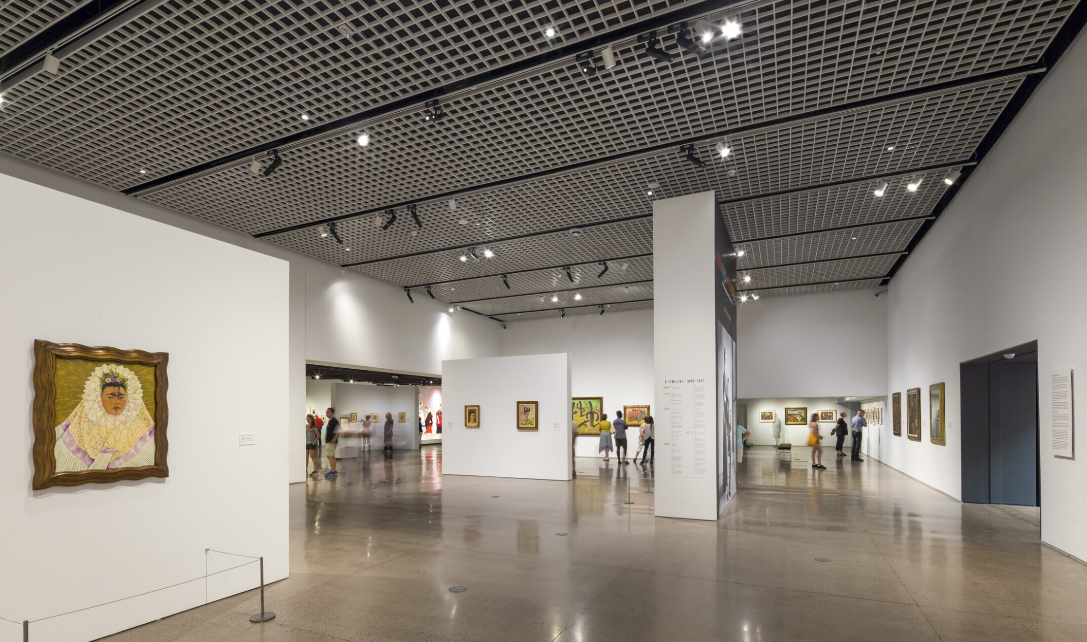 A steel grid with light fixtures hanging off spans the ceiling above a white gallery with paintings on the walls