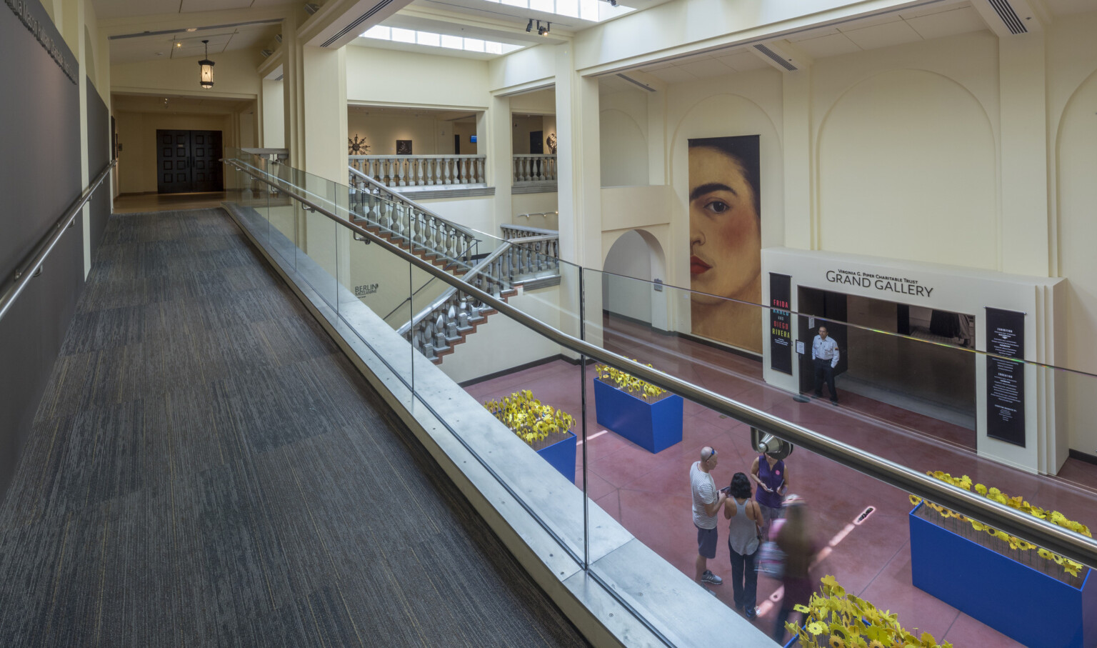 A carpeted walkway overlooks a staircase and an entryway labeled Grand Gallery in cream colored room with skylights
