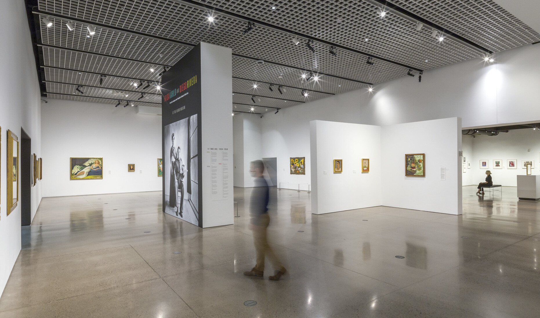A white gallery in the Heard Museum with paintings displayed on the walls and a view of the adjoining gallery